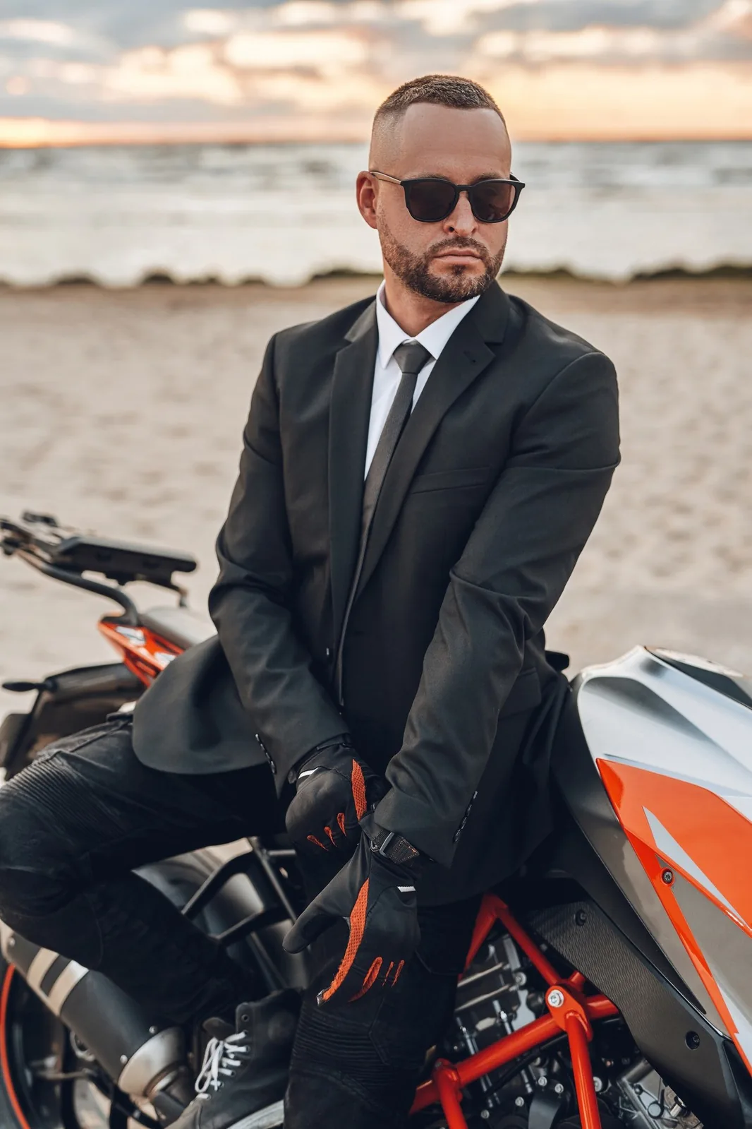 Man in a suit and sunglasses beside a motorcycle near the beach.